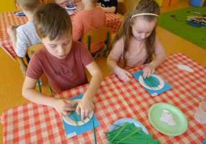 Dwoje dzieci nakleja zielone papierowe łodygi na kartkę papieru.